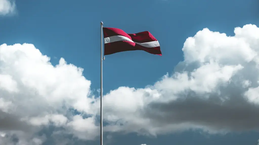 Latvijas valsts karogs plīvo vējā pretī zilām debesīm ar mākoņiem