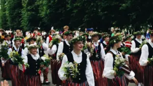Cilvēki tradicionālajos latviešu tautas tērpos piedalās svinīgā gājienā ar ziediem rokās.