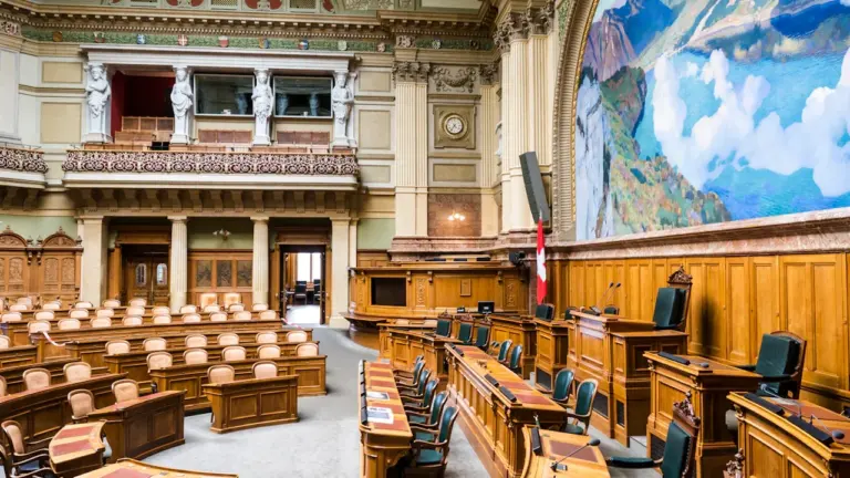 Tukša parlamenta sēžu zāle ar koka galdiem un krēsliem, gatava politiskām diskusijām.